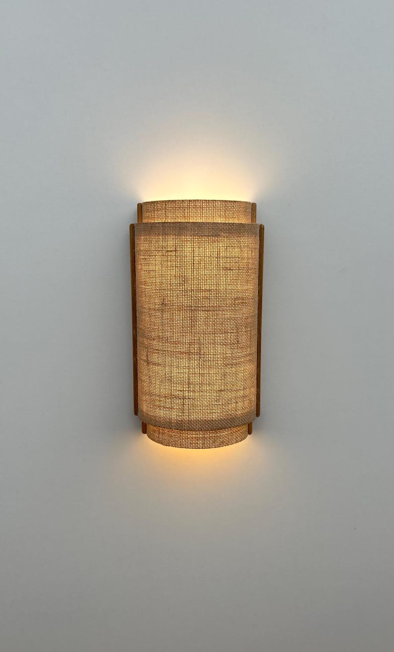 May include: A wall-mounted light fixture with a woven fabric shade. The shade is a light brown color and has a rectangular shape. The light is turned on and illuminating the room.