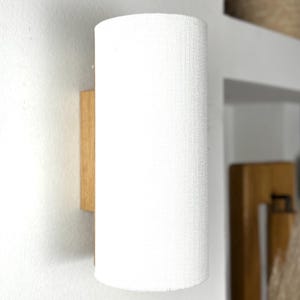 May include: A white cylindrical lampshade with a textured fabric finish, mounted on a wooden wall sconce.