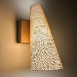 Peut inclure: Un luminaire mural avec un abat-jour cylindrique en tissu tissé. L'abat-jour est de couleur brun clair et a une surface texturée. Le luminaire est monté sur une base en bois.