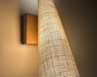 May include: A wall-mounted light fixture with a rectangular wooden base and a cylindrical lampshade. The lampshade is made of a woven, beige fabric, and the light is illuminated from within. The fixture is mounted on a light-colored wall.