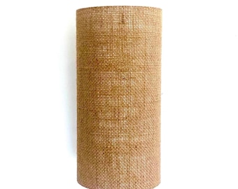 Puede incluir: Una pantalla de lámpara cilíndrica hecha de tela de yute tejida. La pantalla de la lámpara es de color marrón claro y tiene una superficie texturizada. La pantalla de la lámpara está de pie sobre un fondo blanco.