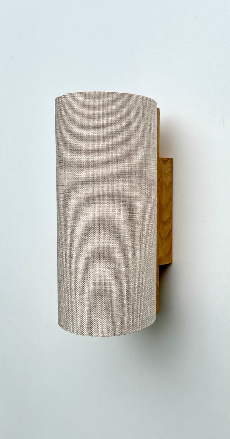 Puede incluir: Aplique de pared cil&iacute;ndrico con pantalla de tela beige y soporte de montaje de madera. La pantalla es de color beige claro y tiene un aspecto texturizado. El soporte de madera es de color marr&oacute;n c&aacute;lido.
