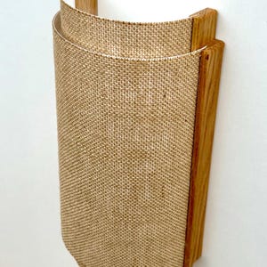 May include: A wall-mounted light fixture with a burlap shade and a wooden frame. The shade is cylindrical and has a natural, rustic look.