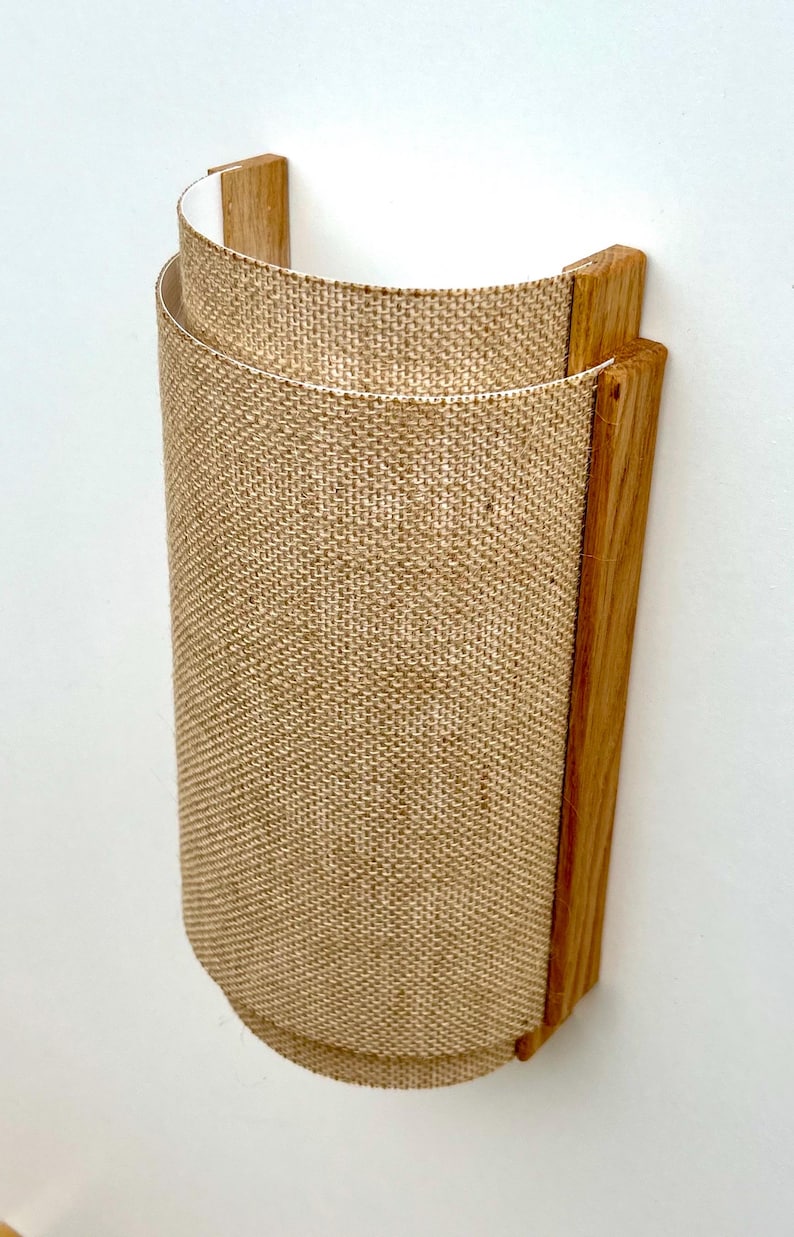 May include: A wall-mounted light fixture with a burlap shade and a wooden frame. The shade is cylindrical and has a natural, rustic look.