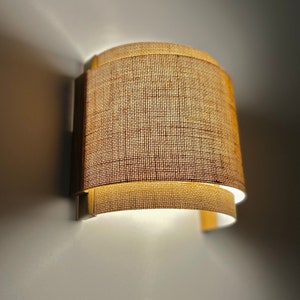 May include: A wall-mounted light fixture with a curved, woven fabric shade. The shade is a light brown color and has a subtle texture.