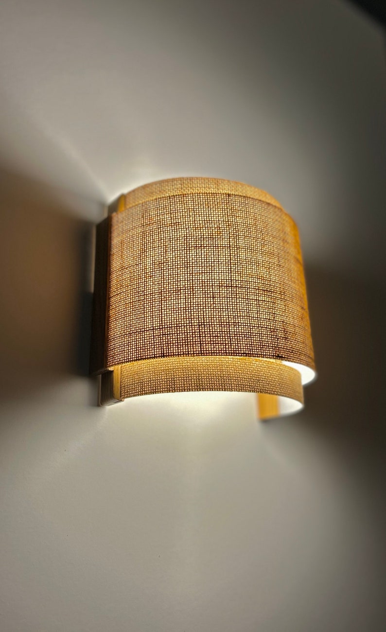 May include: A wall-mounted light fixture with a curved, woven fabric shade. The shade is a light brown color and has a subtle texture.