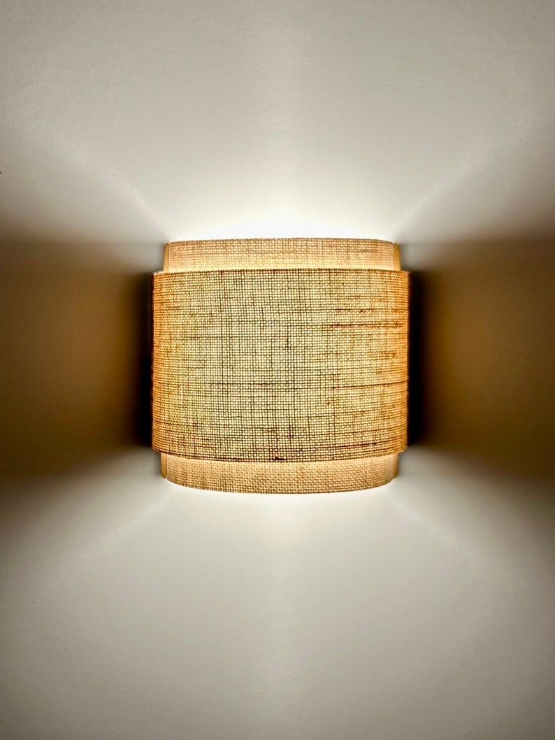 May include: A woven cylindrical lampshade with a light brown color. The lampshade is lit and casting a warm glow.