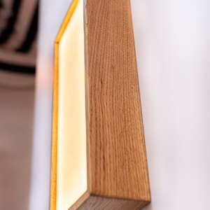 Pu&ograve; includere: Un'applique da parete rettangolare in legno con una luce all'interno. L'applique &egrave; realizzata in legno naturale con una finitura marrone chiaro. La luce &egrave; accesa e illumina la stanza.