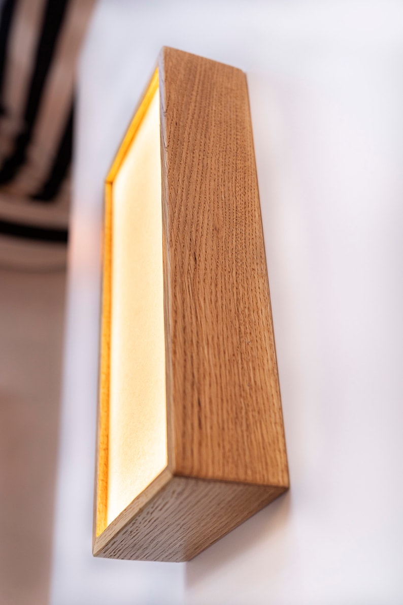 Pu&ograve; includere: Un'applique da parete rettangolare in legno con una luce all'interno. L'applique &egrave; realizzata in legno naturale con una finitura marrone chiaro. La luce &egrave; accesa e illumina la stanza.