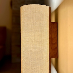 Puede incluir: Una l&aacute;mpara de pared con una pantalla cil&iacute;ndrica de tela beige. La pantalla est&aacute; enmarcada por una parte trasera y una placa de montaje de madera oscura. El accesorio est&aacute; dise&ntilde;ado para montarse en la pared.