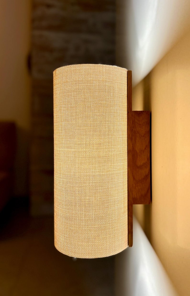 Puede incluir: Una l&aacute;mpara de pared con una pantalla cil&iacute;ndrica de tela beige. La pantalla est&aacute; enmarcada por una parte trasera y una placa de montaje de madera oscura. El accesorio est&aacute; dise&ntilde;ado para montarse en la pared.