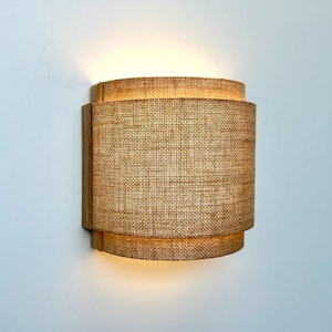 May include: A wall-mounted light fixture with a woven fabric shade. The shade is made of a natural material, like burlap or linen, and has a warm, inviting look. The light fixture is mounted on a wooden base.