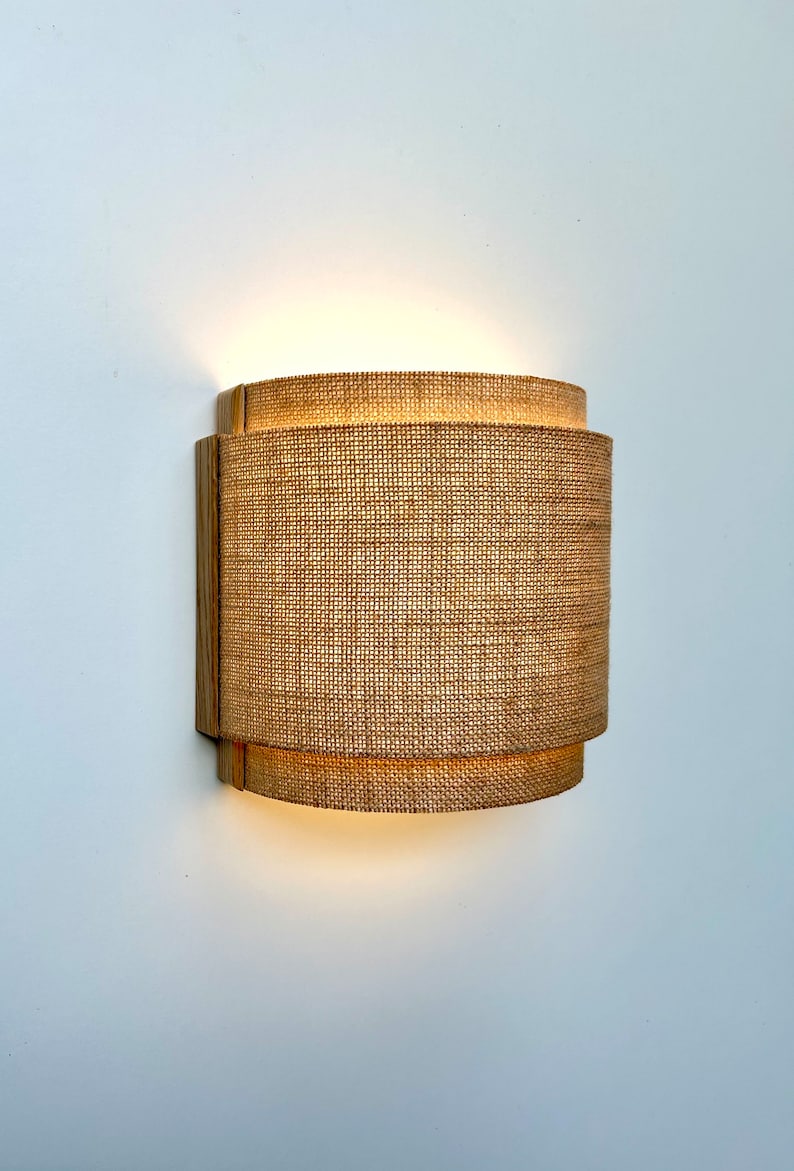 May include: A wall-mounted light fixture with a woven fabric shade. The shade is made of a natural material, like burlap or linen, and has a warm, inviting look. The light fixture is mounted on a wooden base.