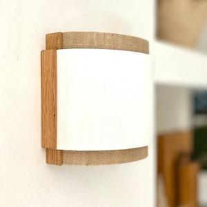 Peut inclure: Un luminaire mural avec un abat-jour en tissu blanc et un cadre en bois naturel. L'abat-jour est bordé de toile de jute.