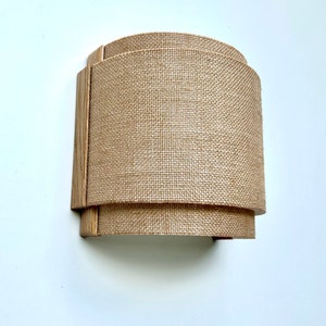 May include: A wall-mounted light fixture with a curved, natural burlap shade and a wooden frame.