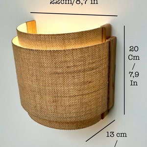May include: A wall-mounted light fixture with a natural burlap shade and wooden frame. The shade is 7.9 inches tall and 5 inches wide. The fixture is 8.7 inches in diameter.