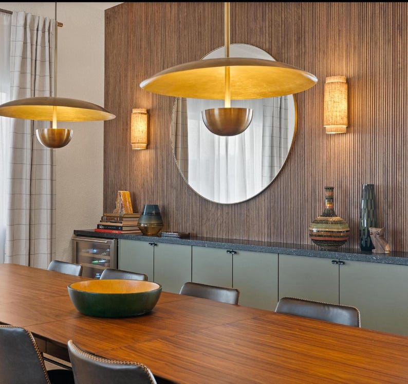 May include: A dining room with a wooden table and chairs. The walls are covered in wood paneling and there are two gold pendant lights hanging from the ceiling. There is a large mirror on the wall and a set of cabinets with green doors.