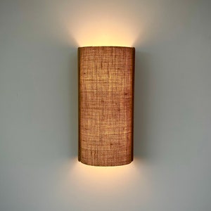 Op de afbeelding: Een wandlamp met een lampenkap van geweven stof. De kap is lichtbruin van kleur en heeft een rechthoekige vorm. Het licht is aan en verlicht de kamer.