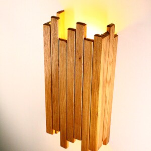 Peut inclure: Un luminaire mural en lattes de bois naturel. Le luminaire est allum&eacute; et diffuse une lumi&egrave;re chaude.