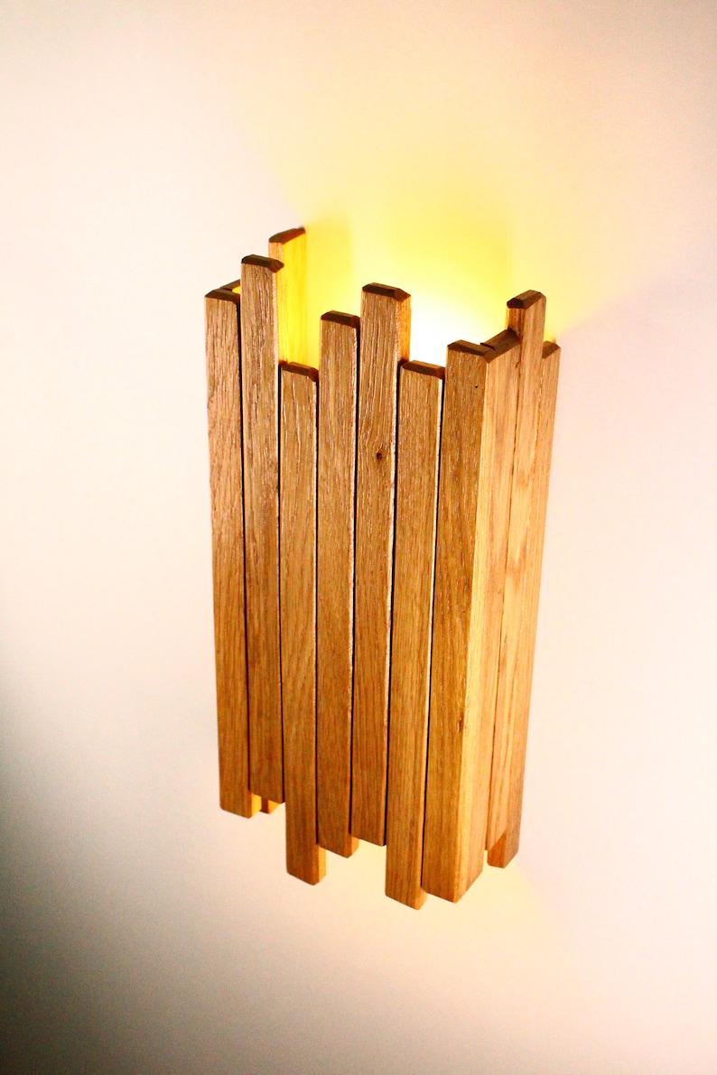 Peut inclure: Un luminaire mural en lattes de bois naturel. Le luminaire est allum&eacute; et diffuse une lumi&egrave;re chaude.