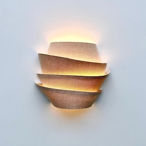 Könnte beinhalten: Moderne Wandleuchte mit geschichtetem Design. Die Leuchte verfügt über eine Reihe von überlappenden, gebogenen Schirmen in einem warmen, natürlichen Farbton, wodurch ein strukturiertes Aussehen entsteht. Die Lichtquelle ist verdeckt und strahlt ein weiches, umgebendes Leuchten aus.