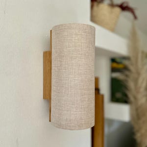 Puede incluir: L&aacute;mpara de pared con una pantalla cil&iacute;ndrica de tela beige. La pantalla est&aacute; unida a una base rectangular de madera. La l&aacute;mpara est&aacute; montada en una pared blanca, con una estanter&iacute;a y objetos decorativos en el fondo.