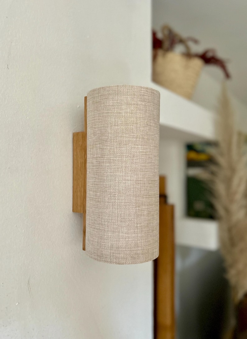 Puede incluir: L&aacute;mpara de pared con una pantalla cil&iacute;ndrica de tela beige. La pantalla est&aacute; unida a una base rectangular de madera. La l&aacute;mpara est&aacute; montada en una pared blanca, con una estanter&iacute;a y objetos decorativos en el fondo.