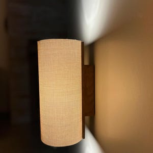 Puede incluir: Una l&aacute;mpara de pared con una pantalla cil&iacute;ndrica de tela beige. La pantalla est&aacute; sujeta por un marco de madera marr&oacute;n oscuro. La luz est&aacute; encendida, proyectando un brillo c&aacute;lido y una sombra en la pared.
