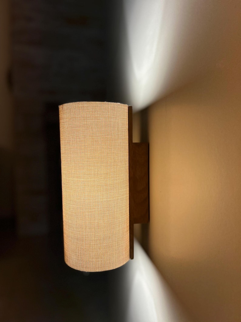 Puede incluir: Una l&aacute;mpara de pared con una pantalla cil&iacute;ndrica de tela beige. La pantalla est&aacute; sujeta por un marco de madera marr&oacute;n oscuro. La luz est&aacute; encendida, proyectando un brillo c&aacute;lido y una sombra en la pared.