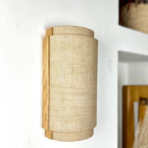 May include: A wall-mounted light fixture with a natural wood frame and a beige linen shade. The shade is cylindrical and has a slightly textured surface.