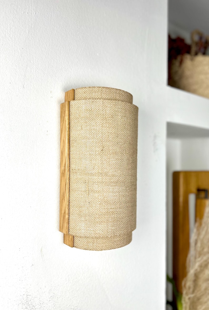 May include: A wall-mounted light fixture with a natural wood frame and a beige linen shade. The shade is cylindrical and has a slightly textured surface.