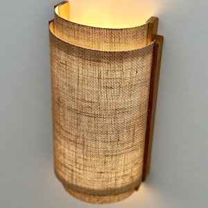 May include: A wall-mounted light fixture with a woven fabric shade and a wooden frame. The light is turned on and emits a warm glow.