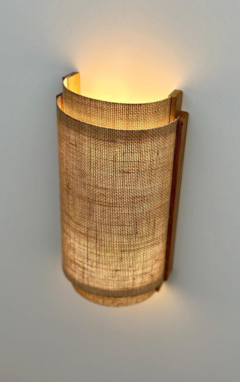 May include: A wall-mounted light fixture with a woven fabric shade and a wooden frame. The light is turned on and emits a warm glow.