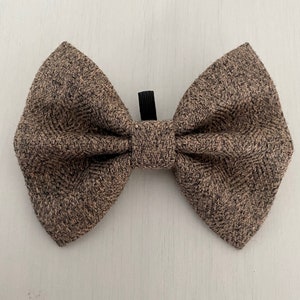 May include: A brown and tan tweed bow tie with a black elastic strap. The bow tie features a traditional shape and a textured pattern. The fabric looks like a woven material, suitable for formal or casual occasions.