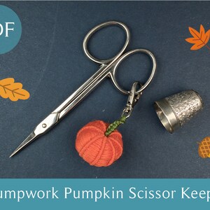 May include: A red and orange pumpkin-shaped stumpwork scissor keeper with a silver thimble and scissors. The scissors have a silver ring with a key chain attached. The background is a dark blue surface with fall-themed leaves and an acorn.