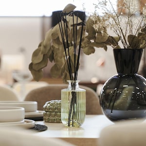 May include: A clear glass diffuser bottle with black reeds and a yellow liquid sits on a white table next to a black ceramic vase with dried flowers.