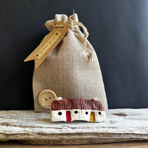 Puede incluir: Una pequeña figura de cabaña de cerámica blanca con techo marrón texturizado, una puerta roja y una puerta amarilla, colocada frente a una bolsa de arpillera beige con una etiqueta marrón que dice Handmade with Love y un botón de madera.