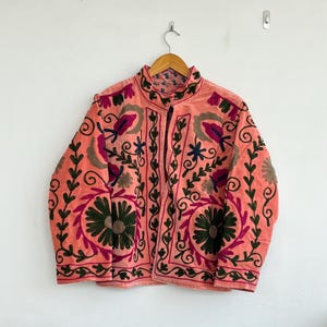 May include: A coral jacket with detailed floral embroidery in green, pink, and purple. The jacket has a mandarin collar and is suspended on a wooden hanger. The embroidery forms a complex pattern across the front and sleeves, suggesting a vintage or bohemian aesthetic.