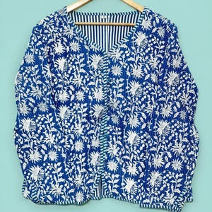 Peut inclure: Veste à imprimé floral bleu et blanc avec un col en V et des manches longues. La veste est suspendue à un cintre en bois sur un fond bleu clair. La veste a un motif à rayures à l'intérieur du col.
