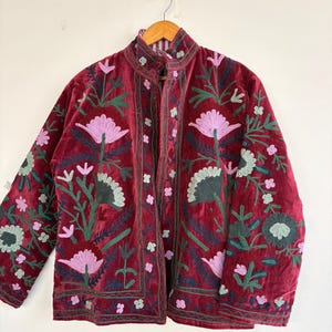 Peut inclure: Une veste en velours bordeaux profond dotée d'un col montant et de manches longues, ornée de broderies florales complexes dans des tons rose clair, vert foncé et sauge pâle. Le vêtement est ouvert sur le devant et suspendu à un cintre en bois.
