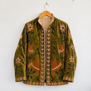 Puede incluir: Una chaqueta de terciopelo verde oliva con mangas largas, cuello alzado y frente abierto. La chaqueta está decorada con patrones florales y motivos de aves bordados en tonos rosa, naranja y verde oscuro, con un forro interior a rayas.