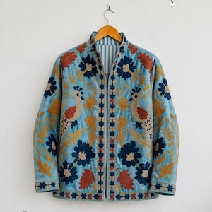 Könnte beinhalten: Eine hellblaue, gesteppte Jacke mit aufwendigen Stickereien aus dunkelblauen Blumen, goldgelbem Blattwerk und zwei Vögeln mit rotbraunen Flügeln. Die Jacke hat einen Stehkragen, lange Ärmel und eine vertikale Vorderöffnung mit gestreiftem Futter.