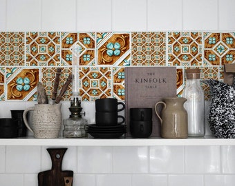 May include: A white shelf with various kitchen items, including a book titled "The Kinfolk Table", a glass pitcher, a black and white speckled pitcher, a ceramic pitcher, a glass oil lamp, and black coffee cups. The shelf is against a white tiled wall with a decorative tile backsplash featuring a repeating pattern of blue and gold.