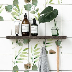 Könnte beinhalten: Ein weiß gefliestes Badezimmer mit einem Holzregal, auf dem drei Flaschen Duschgel und Handcreme stehen. Die Flaschen sind mit "meraki" beschriftet. Ein Handtuch hängt an einem Haken und eine Holzbürste steht auf dem Regal. Die Fliesen haben ein Aquarelldesign mit grünen Blättern.