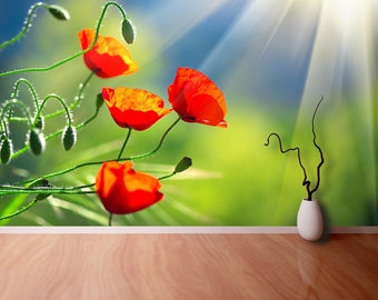 May include: A close-up of red poppy flowers with green stems and leaves against a blurred background of blue sky and green grass. The sun shines brightly in the background.