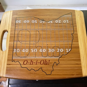 Könnte beinhalten: Ein Schneidebrett aus Holz in Form des Bundesstaates Ohio mit einem Footballfeld-Design und dem aufgedruckten Text "O-h-i-Oh!"