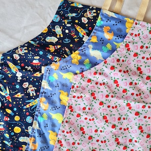 May include: Three colourful fabric aprons with different patterns. The first apron is navy blue with a space theme, the second is light blue with a dinosaur theme, and the third is pink with a strawberry theme.