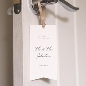 Puede incluir: Una etiqueta rectangular blanca para puerta con el texto "THIS ROOM IS RESERVED FOR" y "Mr & Mrs Johnstone BRIDAL SUITE" en escritura elegante. La etiqueta cuelga de una manija plateada con una cinta beige.