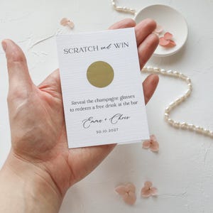 Peut inclure: Une carte à gratter rectangulaire blanche avec une zone à gratter circulaire dorée. Le texte indique SCRATCH and WIN, Reveal the champagne glasses to redeem a free drink at the bar, Emma + Chris, et la date 30.10.2027 en polices noire.