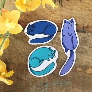 K&ouml;nnte beinhalten: Drei Katzenaufkleber mit verschiedenen Posen. Die Katze links ist blau und zusammengerollt. Die Katze in der Mitte ist t&uuml;rkis und zusammengerollt. Die Katze rechts ist lila und steht auf.